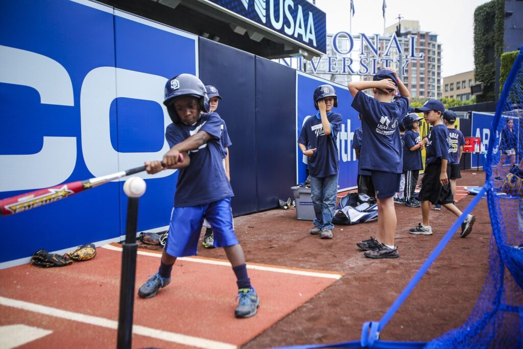 Young player in a batting helmet hitting off a tee while other kids and coaches wait their turn at a group youth baseball clinic