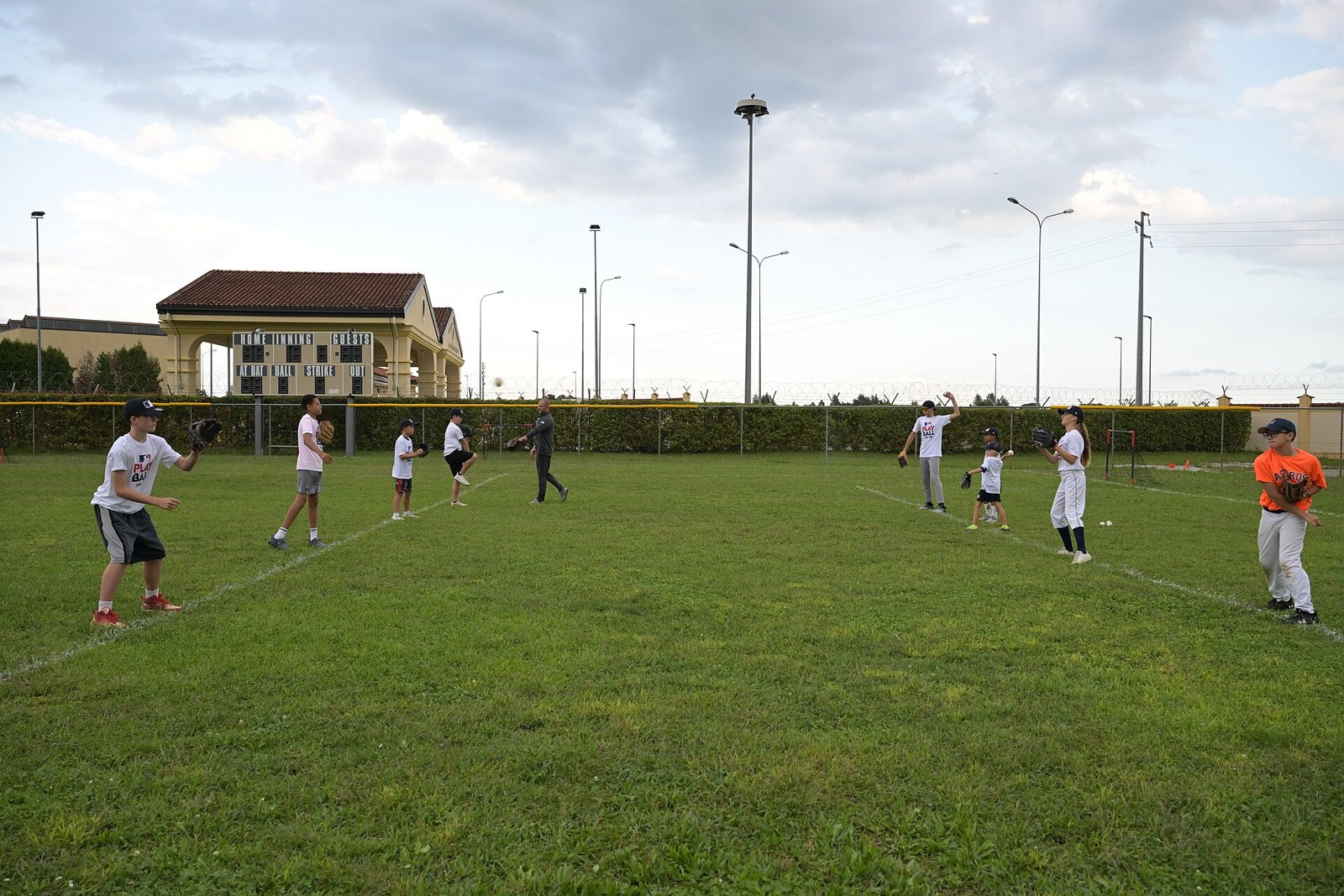 Youth players spread across a grass field running throwing drills, similar to the stations at a select baseball tryout