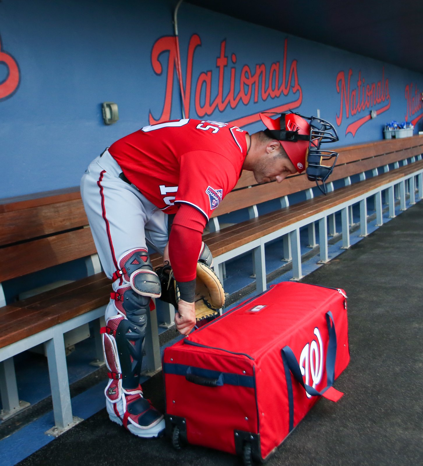 Catcher in leg guards pulling gear out of a wheeled equipment bag in the dugout
