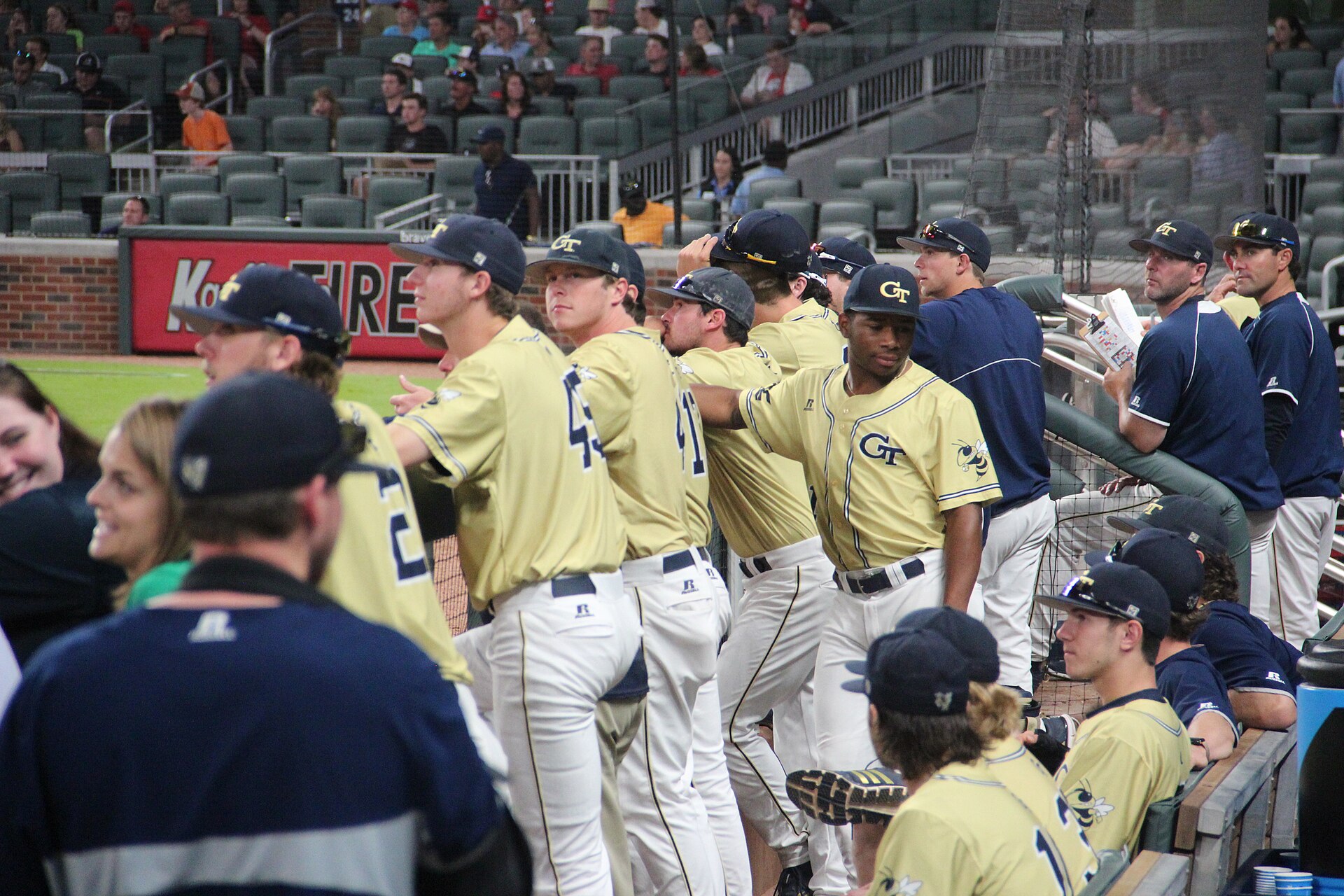 College baseball players standing in the dugout during a game, where cold weather baseball gear earns its keep between innings