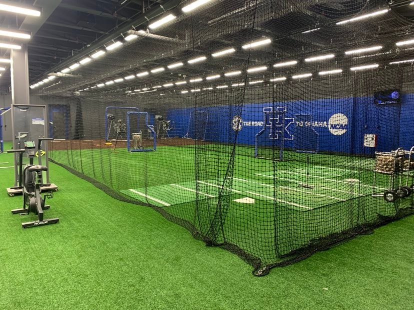 Rows of netted indoor batting cages on turf with pitching machines, the setup most indoor baseball training facilities in North Texas use