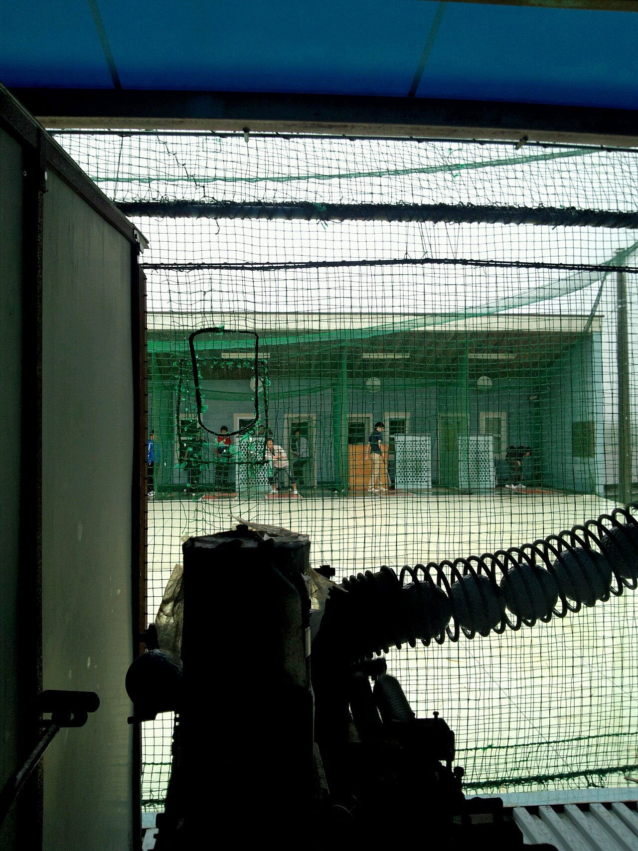 View from inside an indoor batting cage toward a pitching machine, similar to the token machine lanes at indoor baseball training facilities in North Texas
