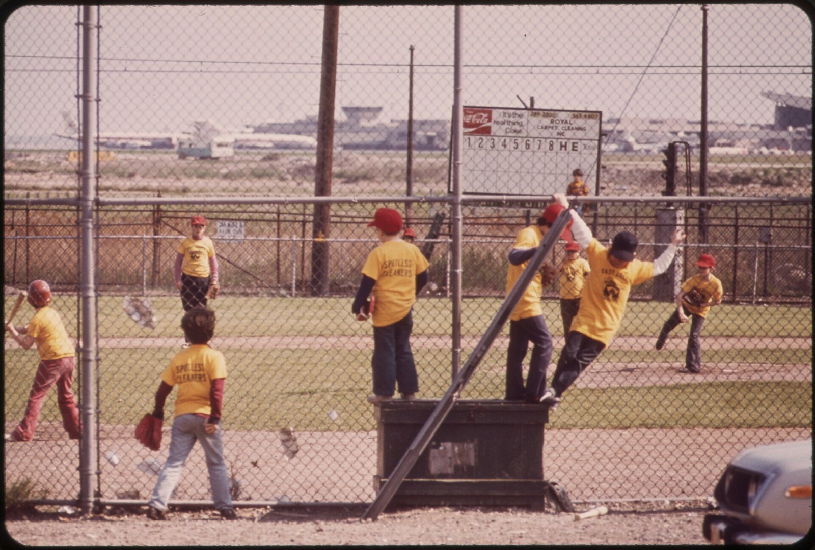 Little League players in yellow team shirts along the backstop fence during a 1973 game near Logan Airport in Boston, with a batter holding a bat at left