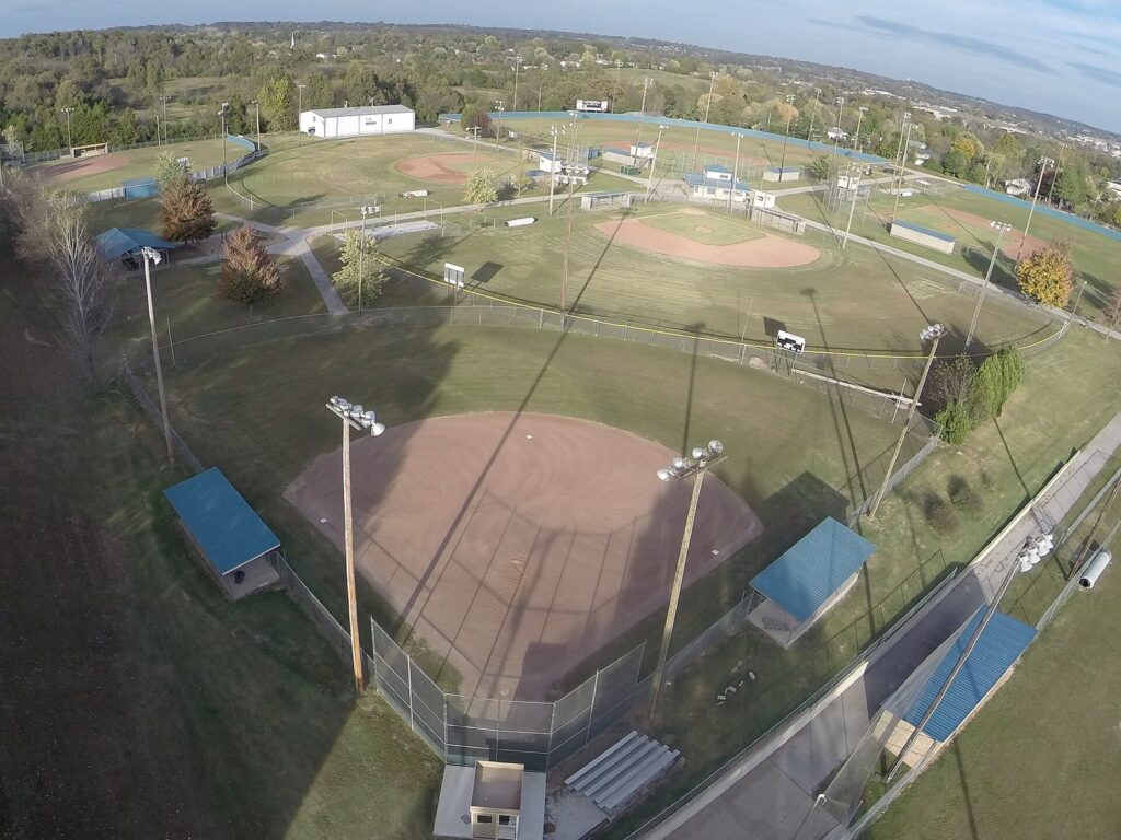 Aerial view of a multi-field youth baseball and softball complex with lights and covered dugouts, the kind of venue that hosts tournament weekends