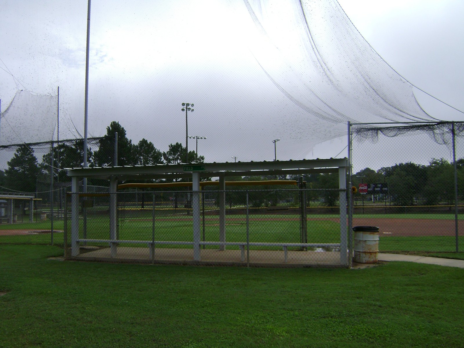 Empty dugout and backstop netting at a youth baseball complex on an overcast day