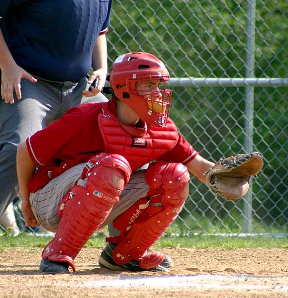 Young catcher crouching in red helmet, chest protector and leg guards with mitt up as a target